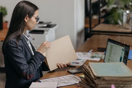 Administratoare lucrând la dosare, cu un tabel deschis pe laptop și calculatorul de birou alături