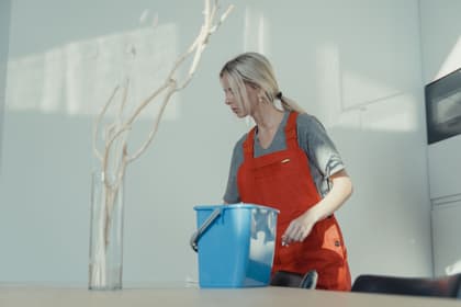 Agent de curățenie în șorț de lucru, cu o găleată, ștergând o masă
