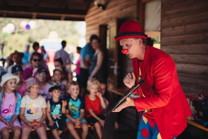 Animator costumat în clovn jucând un spectacol pentru un grup de copii, lângă o cabană de lemn