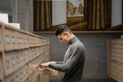 Arhivist căutând printre fișele dintr-un sertar, într-o sală cu fișiere de lemn