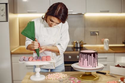 Cofetăreasă umplând macarons cu cremă din poș, lângă un tort