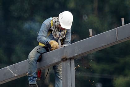Montator de structuri metalice lucrând cu polizorul pe o grindă de oțel, la înălțime