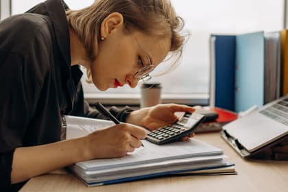 Contabilă completând un registru, cu calculatorul de birou în mână
