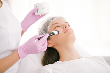 Cosmeticiană aplicând cu pensula un tratament pe tenul unei cliente, într-un cabinet cosmetic