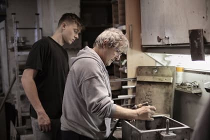 Meșter arătându-i unui ucenic cum se lucrează o piesă, la bancul de lucru al unui atelier