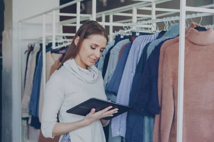 Lucrătoare comercială verificând marfa pe o tabletă, între rafturile cu haine ale unui magazin