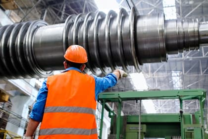 Muncitor cu cască și vestă reflectorizantă lângă rotorul unei turbine cu abur, într-o hală industrială