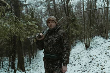 Paznic de vânătoare în haine de camuflaj, cu arma pe umăr, într-o pădure de conifere înzăpezită