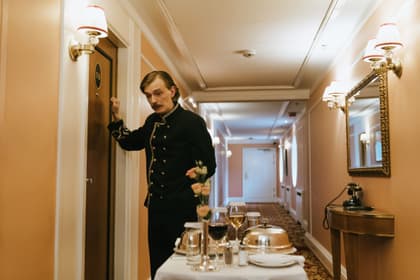 Angajat de hotel în uniformă bătând la ușa unei camere, cu un cărucior de servire pe hol