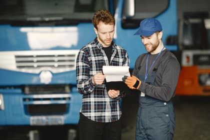 Tehnician transporturi verificând documentele unui transport împreună cu un coleg, în fața camioanelor